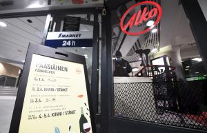 En del Alko-affärer har förkortade öppettider under torsdag och lördag. Till vänster syns en skylt där öppettider står listade, till vänster en skylt med texten alko. I bakgrunden syns en person med en kundvagn.