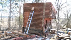 En röd stockbyggnad står på en gård med högar av material och bråte. Byggnaden saknar tak.