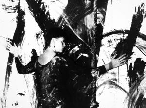Mustavalkoinen kuva, jossa Marikki Hakola maalaa valkoiselle seinälle vartalollaan mustia jälkiä.