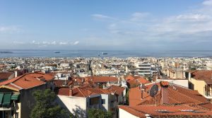 Vy över takåsarna i Thessaloniki där det syns tegelpannor på ljusa hus och på redden ligger några lastfartyg.