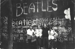 Neljä nuorta aikuista poseeraa iloisesti seinän edessä. Seinään on kirjoitettu The Beatles -yhtyeen jäsenten nimiä ja piirretty rauhanmerkkejä.