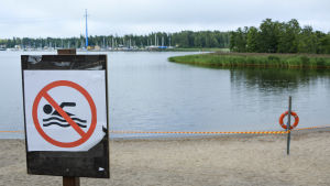 För tillfället råder badförbud vid badstranden vid Smulterö. Skylt som visar att det är förbjudet att bada. I bakgrunden syns en avspärrad badstrand.