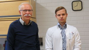 Helge Heinonen och Samuel Henriksson, som är andra sommaren i rad på jobb vid Ingå hälsocentral. Unga läkare som rekommenderar hälsocentralen till andra unga läkare är den bästa marknadsföringen, konstaterar Helge Heinonen. En man med grått hår och mustasch och en yngre man med vit läkarrock inne i ett mottagningsrum. De tittar in i kameran.