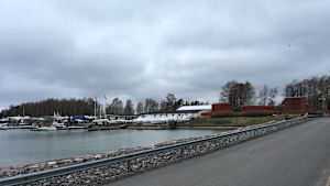 Björkholmen just nu, sedd från Drumsö. Bron till Björkholmen från Drumsö. Himlen är mulen, havet grått. På bilden syns båtar uppe på land och röda magasinbyggnader.