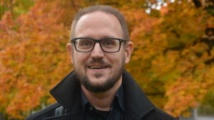 Forskare Michael Kull har forskat i vad som gör staden Närpes attraktiv.. Man i glasögon och skägg ler mot kameran med höstlov i bakgrunden.