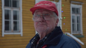 Ingmar Holmström är pigg, men upp på taken klättrar han inte längre. Sonen brukar hjälpa Ingmar Holmström med diverse nödlösningar. En äldre man med röd keps står utanför ett gult trähus. Vinter och snö.
