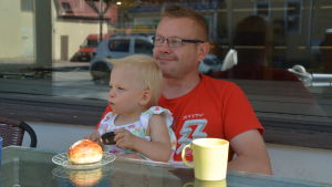 Janne Ylönen med gudbarnet i famnen tittar efter bilar på Lappträskvägen. Janne Ylönen sitter vid ett bord och tittar ut mot vägen, han har sitt gudbarn i famnen.