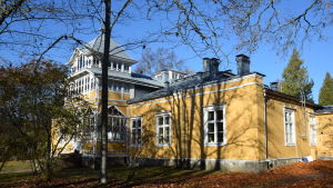 Ett av Mikaelskolans skolhus är en gammal gul träbyggnad. Ett gult gammalt hus skymtar bakom några träd. På markens ligger höstlöv.
