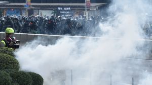 Kravallpoliser har skjutit tusentals tårgaskanistrar mot demonstranterna. De anklagar polisen för att ha gjort sig skyldig till kriminell brutalitet. Kravallpoliser skjuter tårgas mot demonstranter. Ett moln av rök och i bakgrunden en skylt med texten "Warning tear smoke". Polisen anklagas för kriminell brutalitet.
