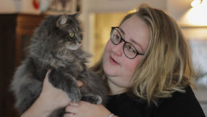 En vit kvinna med blont hår och svarta glasögon ler. och håller upp en långhårig, grå katt.
