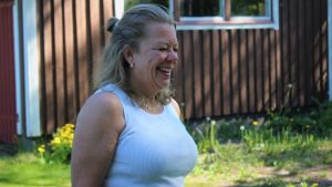 Det tar mycket att vara fosterförälder, men det ger också mycket, konstaterar Nina Träsk. Att se Hanna utvecklas och må bra är belöningen. Kvinna i medelåldern klädd i ett vitt linne i somrig miljö ser bort från kameran och skarttar,