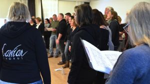 På första övningen sjöngs bland annat Somewhere over the rainbow från Trollkarlen från Oz och Willkommen från Cabaret. Noter i förgrunden, körsångare sjunger. Vi ser att det står Cawiar på ryggen på en sångares munkjacka.
