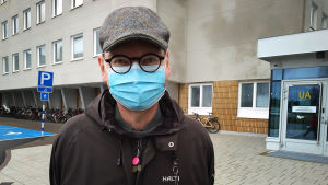 Ledande överläkare Heikki Kaukoranta är skeptisk till beslutet att ta emot utbytesstuderande. En man med runda svarta glasögon, rutig keps och munskydd står utanför sjukhuset.