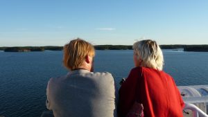 En man i grå tröja och en kvinna i röd tröja står på ett fartygsdäck och tittar ut över en stilla skärgård.