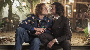 Elton John (Taron Egerton) förälskad i den blivande managern John Reid (Richard Madden). Elton John (Taron Egerton) och John Reid (Richard Madden) sitter tätt tillsammans på en trappa och ler.
