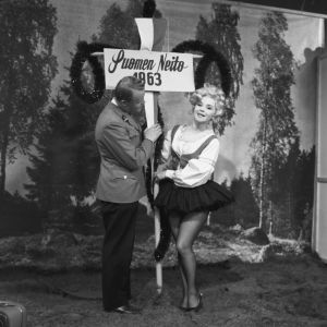 Taikuri Reijo Salminen ja näyttelijä Marjatta Kallio Iskelmäkarusellissa 1963. Kallio on pukeutunut lyhythelmaiseeen kansallispukumuunnelmaan ja kantaa Suomen Neito 1963 -kylttiä.