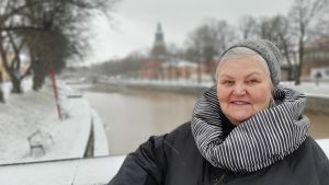 Lena Sarlin bjuder gärna hem turister, så att de ska få uppleva skärgården och få smaka på äpplen och hallon i hennes trädgård. Lena Sarlin, en dam med grånande hur, grå mössa och en svartvitrutig halsduk står vid ett snöigt broräcke. I bakgrunden syns Aura å och Åbo Domkyrka.