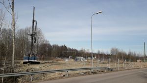 Så här ser det ut just nu, i början av april 2020. Grundläggningsarbeten, man förbereder en tomt för höghusbyggen vid Brännmalmsvägen i Sjundeå. En stor lyftkran eller lift och på håll kan man skönja lastbilar. Annars tom tomt utan träd invid en asfalterad väg.