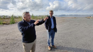 Byggföretagaren Jan-Erik Chydenius är glad över att byggarbetena inletts. Här tillsammans med tjänstemannen Jukka Takala från Hangö stad. Jan-Erik Chydenius står och visar något med händerna. Jukka Takala står bredvid och tittar. I bakgrunden skymtar det öppna havet.