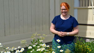 Johanna Lönnfors. En kvinna står omringad av prästkrage blommor framför ett trähus