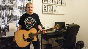Tom Håkans skapar ofta musik hemma framför datorn. Tom Håkans står inomhus med gitarr i handen. I bakgrund finns en dator på ett skrivbord, en synth och på väggen finns tavlor med cd-skivor.