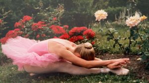 Mari Rosendahl balettipuku ja -tossut yllään nurmikolla sirossa istunnassa ylävartalo suorien jalkojen yli taivutettuna. Taustalla ruusuistutuksia.