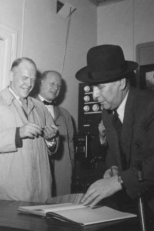 Invigning av UKV-sändaren i Ekenäs år 1.10.1954. Mannen som skriver sitt namn i gästbokgen är sannolikt Yles tekniska chef Paavo Velander. Till vänster chefen för Yles svenska verksamhet Ragnar Ölander och Ekenäs stadsfullmäktiges ordförande Sven Högström. Tre män står omkring en uppslagen bok.