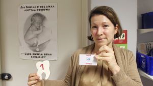 Patricia Clayhills-Siljama visar upp en namnskylt. Vårdarna är anonyma, så istället för ett namn står det bara hälsovårdare på lappen. patricia står framför en skylt som uppmanar till att använda kondom, hon visar upp en namnskylt där det bara står vårdare