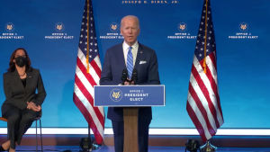 Joe Biden blir USA:s 47:e president. En man i blå kostym och blå slips står vid ett podium, bakom honom syns två amerikanska flaggor. Till vänster en kvinna i granitgrå kostym och svart munskydd. Hon sitter på en stol.