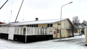 Restaurangen har en terrass mot havet. Ett gammalt gult magasin med en uteservering omgärdad av staket och fönster.
