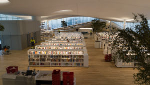 Helsingfors centrumbibliotek Ode. Helsingin keskustakirjasto Oodin 3. kerros. Avarassa tilassa on kumpuileva valkonen katto, puisella parkettilattialla on useita valkoisia kirjahyllyjä. Punaisia muuttolaatikoita on muutamassa pinossa.