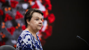 Kirsi Varhila. Kirsi Varhila sitter framför en mikrofon med ett förstorat coronavirus i bakgrunden.