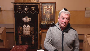 Harry Serlo i synagogan i Åbo. Harry Serlo, en man med gråsvart munkjacka och en kippa på huvudet, står framför läspulpeten och altaret i den lilla salen i synagogan i Åbo.