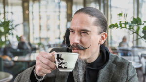 Viiksekäs Juhana Helmenkalastaja nauttii kahvia viiksikupista. 