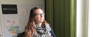 En kvinna ser till sin vänster. Hon står inne på ett kontor. Solen lyser på hennes ansikte från ett fönster. Hon ser seriös ut.