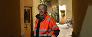 Ben Mattsson. Ben Mattsson i orange kläder står i en portgång med en julgran i bakgrunden.