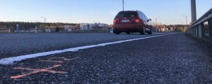 Korsningen också här sedd från riksväg 25 men nu västerifrån, från Ekenäs- och Hangöhållet. På söndagskvällen (5.5) var trafiken inte så livlig. På asfalten har det målats märken på grund av undersökningarna. En T-korsning. En röd personbil som kör mot korsningen syns bakifrån. Det syns också trafikmärken, vägräcke och affärsbyggnader.