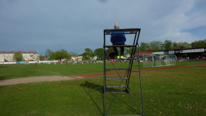 Eino Fredriksson sitter vid korta ändan av planen. Eino Fredriksson sitter på en ställning för att se fotbollsmatchen bra då han syntolkar. Det är ganska svårt att se bortre planen.