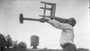 En man lyfter en stol vågrätt med sina bara tänder, samt en slägga med sin arm, samtidigt. Bilden är tagen 1931 i Närpes, Kalaks.