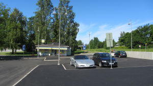 Macken ska byggas bakom kiosken på bilden. En parkering där några bilar står parkerade. I bakgrunden syns en kiosk.