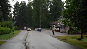 I somras körde mopedister bland annat längs Sockenvägen i Tenala i hög hastighet. En väg där man ser två bilar köra och en kvinna gå med sin hund. På ena sidan av vägen syns några byggnader.