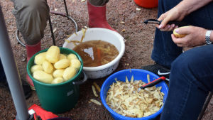 Marthorna skalar potatis i ungefär två timmar. Några mänskor sitter och skalar potatis. På marken syns två handfat med potatisskal och ett ämbar med färdigskalad potatis.