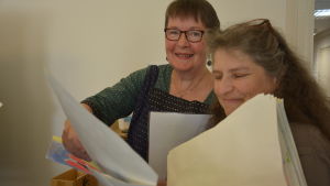 Artesan Sari Kippilä (till höger på bilden) hjälpte till med kursens konstnärliga del. Till vänster Marianne Jokinen. Två kvinnor bläddrar bland teckningar.