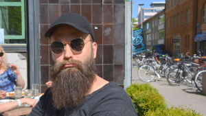 Mikko Manninen Mikko Manninen is sitting outside and wearing round sunglasses and a black cap. He has a long beard.