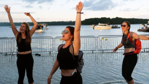 Inget dåligt ställe att dansa på - invid strandbrynet på Weekend i Helsingfors. Tre kvinnliga besökare av Weekend-festivalen i Helsingfors sträcker upp sina händer i luften samtidigt som de dansar nära strandkanten. Bakom syns havet och båtar som lagt till invid festivalområdet.