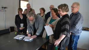 Veijo Lücke (FS) undertecknar partiernas programförklaring. Till vänster om honom Lea Räisänen (G). Från vänster: Tomy Wass (VF), Eva-Stina Hellbom (SDP), Kalevi Kallonen (C), Ghita Edmark (SFP), Janina Kaita (Saml) och Bo Vilander (SDP). Flera personer samlade för att skriva under ett papper.