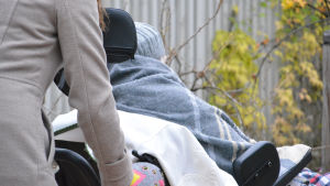 Hur mycket eller lite ska taxichauffören få hjälpa en färdtjänstklient? Och ska hen få betalt för det? Kvinna skjuter på rullstol där det sitter en äldre person.