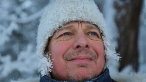 Lemmenjoella pakkanen kuorrutti dokumentaristin kuuraukoksi. Miehen kasvot lumisessa maisemassa. Pipon alta pilkistävät hiukset ovat valkeassa kuurassa ja myös pipoa peittää hentoinen kuura.