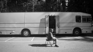 Mies istuu parkkipaikalla bussin vieressä.
