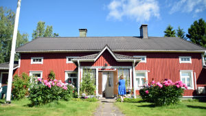 Caféet är inrett i den gamla släktgården. En gammal bondgård med vita knutar med en grönskande trädgård framför. En kvinna i blå klänning syns gå mot husets ingång.